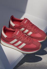 Paire de baskets rouges Adidas avec des bandes blanches et des semelles gomme, placées sur une surface blanche inclinée sous un éclairage doux.