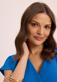 Niebieska bluzka z krótkimi rękawami. Gładka tekstura, dekolt w kształcie litery V i efekt warstwowy. Złota bransoleta na nadgarstku i neutralne tło.