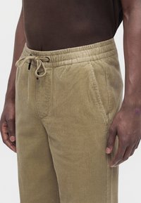 Persona che indossa pantaloni di velluto beige con vita elastica e coulisse, abbinati a una camicia marrone scuro, in piedi davanti a uno sfondo semplice.