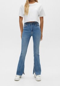 Flared blue jeans in light denim with side slits, high waist, and a smooth texture. Paired with a cropped white T-shirt and white shoes.