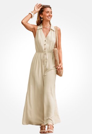 Kobieta uśmiechająca się, ubrana w bezrękawnikowy beżowy kombinezon z guzikami, kolczyki w kształcie rozgwiazdy, naszyjnik z muszli, słomianą torebkę na ramię i beżowe sandały.