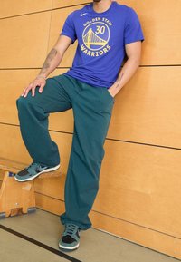 Modré tričko s logem "Golden State Warriors", zelené široké kalhoty a černé/modré tenisky. Viditelné dřevěné pozadí a sportovní podlaha.