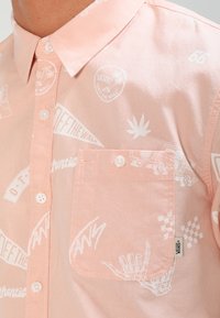 Hellrosa Hemd mit Knopfleiste, das weiße grafische Drucke, darunter Logos und Muster, sowie eine Brusttasche und einen regulären Kragen aufweist.