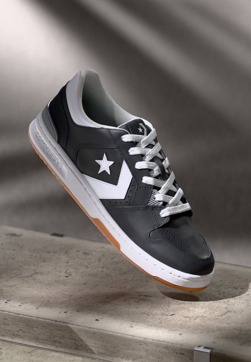 Black leather sneaker with white accents, featuring a star logo, rubber sole with gum bottom, and white laces. Simple, low-profile design.