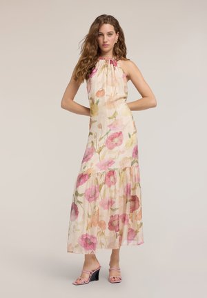 Mujer con cabello largo y ondulado lleva un vestido maxi sin mangas con estampado floral en tonos rosa y beige y sandalias rosas con tacón, de pie con las manos en las caderas.