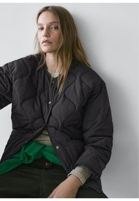 Ung kvinde med langt hår iført en sort quiltet jakke over en grå top og en grøn sweater, der sidder op ad en ensfarvet hvid væg.