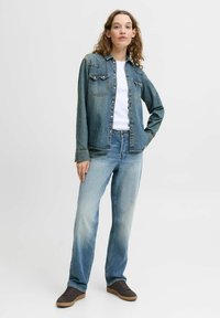 Giacca di jeans in blu sbiadito con due tasche sul petto, chiusura con bottoni e dettagli cuciti, abbinata a jeans blu chiaro e sneakers marroni.