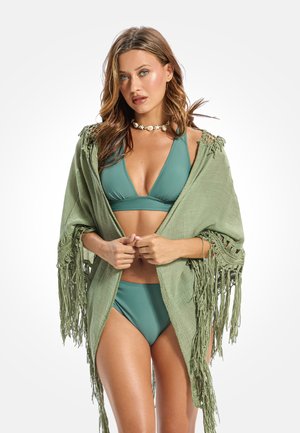 Kobieta w bikini koloru morska zieleń i zielonym szalem z frędzlami, trzymająca brzegi szala, z naszyjnikiem z muszli na białym tle.