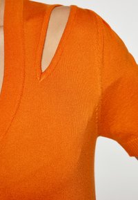Top tricotat portocalie cu decolteu adânc în V și detalii cu bretele tăiate. Textură netedă, cu un design ajustat și mâneci scurte.