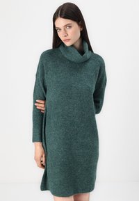 Knit green turtleneck dress with long sleeves, a relaxed fit, and side slits; texture is soft and slightly chunky.