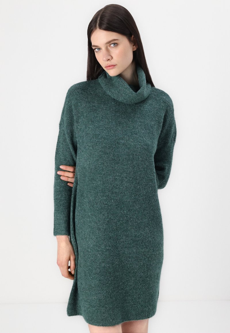 Vestito a collo alto verde in maglia, con maniche lunghe, vestibilità comoda e spacchi laterali; la texture è morbida e leggermente spessa.
