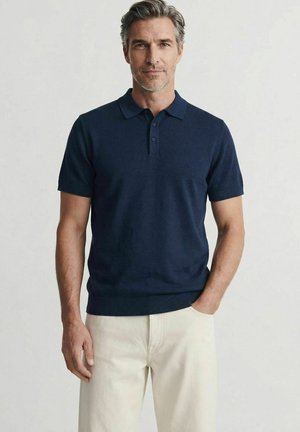 Mann mit grauen Haaren, der ein marineblaues Poloshirt mit kurzen Ärmeln und cremefarbene Hosen trägt, steht mit einer Hand in der Tasche.