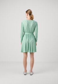 Rochie verde mentă cu talie ajustată, mâneci lungi și un model în diamant. Materialul are o textură netedă. Asortată cu pantofi argintii.