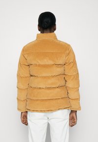 Chaqueta de plumas de pana en color mostaza, con cuello alto, secciones acolchadas horizontales divididas y superficie texturizada.