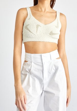 Kobieta w białym, teksturowanym crop topie z wzorami logo Nike oraz białych, wysokich spodniach z wycięciami w talii.