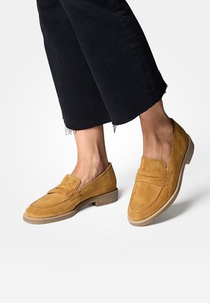 Mocassins en daim marron avec détails cousus, semelle plate et accent minimaliste avec une boucle. Portés avec un pantalon noir raccourci.