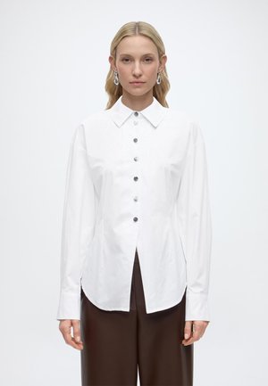 Kobieta o blond włosach ubrana w białą, zapinaną na guziki bluzkę z długimi rękawami oraz brązowe spodnie z wysokim stanem, stoi na jednolitym tle.