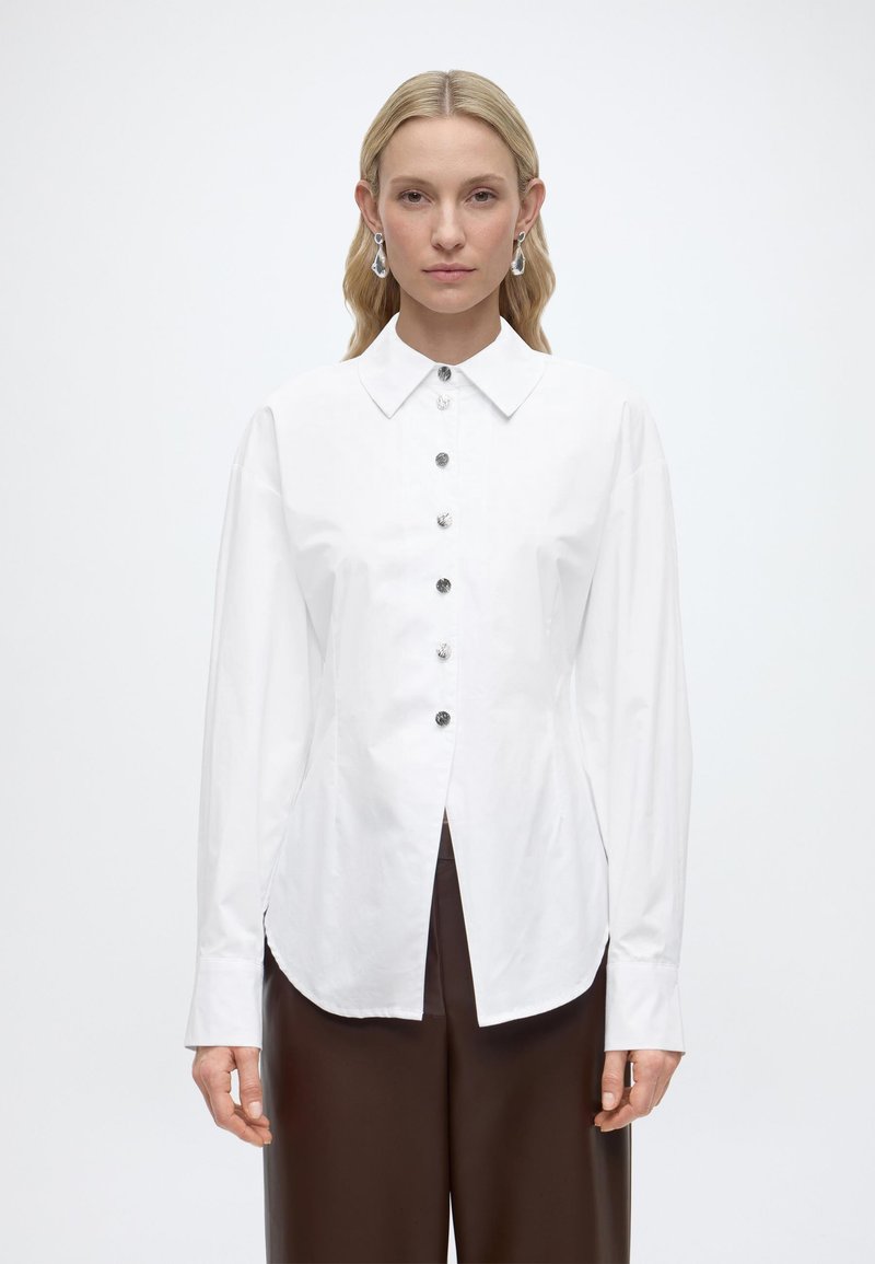 Femme aux cheveux blonds portant un chemisier blanc à boutons avec des manches longues et un pantalon marron taille haute, debout devant un fond uni.