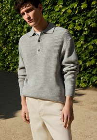 Grau gerippter Strickpullover mit Kragen und drei Knöpfen, kombiniert mit hellen Hosen. Grüne Blätter im Hintergrund.