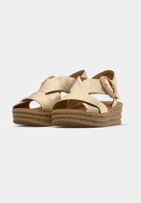 Paire de sandales compensées beiges avec des brides en cuir croisées et une boucle ronde sur une semelle texturée sur fond gris clair.