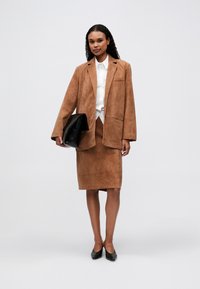 Vrouw in een camel kleurige suède blazer en rok, met een witte blouse, zwarte puntige schoenen, die een grote zwarte clutches draagt, staand tegen een effen achtergrond.