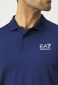 Ναυτικό μπλε πόλ�ο μπλούζα με υφασμένο ύφασμα, τρία μαύρα κουμπιά και λευκό κεντημένο λογότυπο με "EA7 Emporio Armani" στο στήθος.