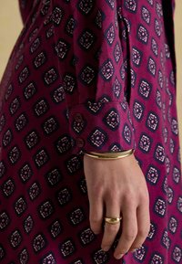 Dlhý rukáv šaty s magentovým základom a geometrickým vzorom v námorníckej modrej a bielej farbe. Obsahujú gombíkový manžet a kovový náramok na zápästí.