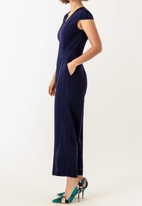 Tmavě modrý overal s přiléhavým živůtkem, krátkými rukávy a bočními kapsami. Kombinován s vícero barevnými vysokými podpatky s květinovým vzorem.