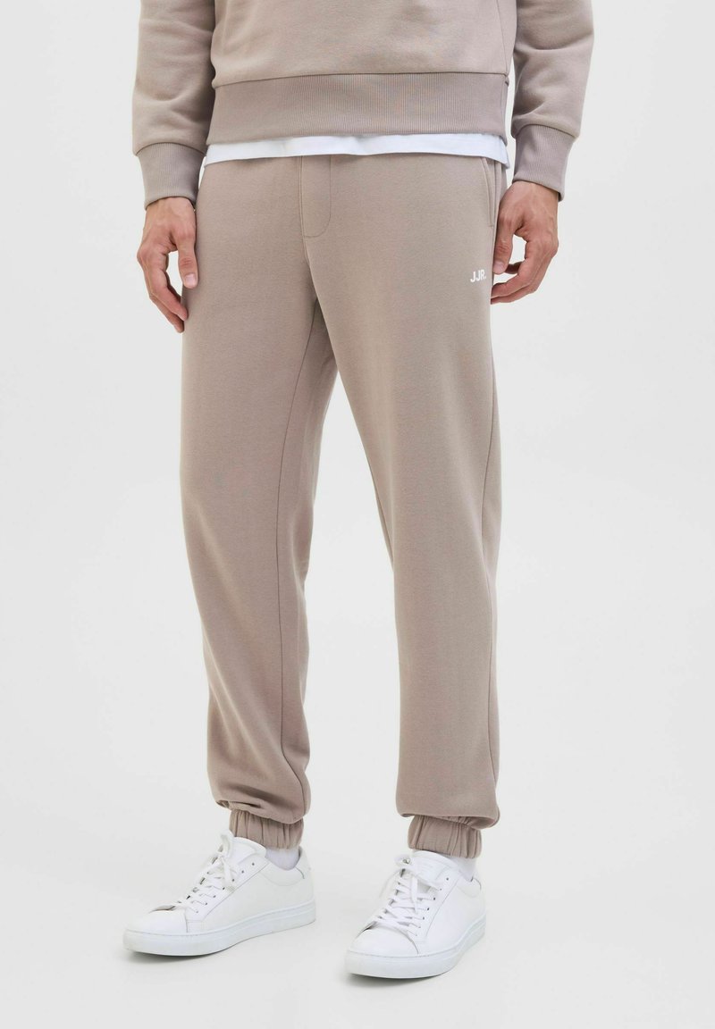 Persoon draagt beige joggerbroeken met elastische boorden en witte sneakers, staand tegen een effen witte achtergrond.