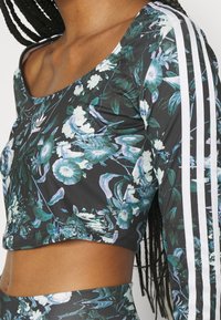 Sort blomster crop-top har et livligt botanisk print med grønne og lilla accenter. Tre hvide striber løber langs ærmerne.