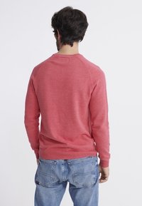 Homme portant un pull rouge à manches longues et un jean bleu, debout de dos face au spectateur, sur un fond blanc uni.