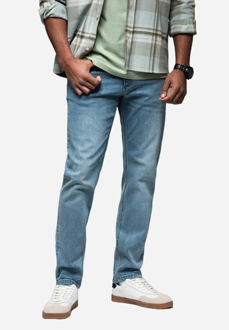 Jeans de color azul claro con un corte relajado, combinados con zapatillas blancas. El atuendo incluye una camisa de cuadros abotonada sobre una camiseta verde.