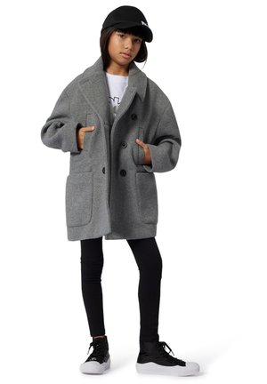 Manteau en laine grise à coupe oversized, avec de grandes poches avant et des boutons double boutonnage. Porté sur un t-shirt blanc et des leggings noirs. Casquette noire et baskets.