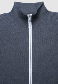 Maglione grigio a coste con colletto alto. Presenta una zip bianca centrale e una striscia bianca a contrasto lungo la linea della zip. Texture morbida.