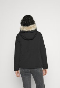 Person med lockigt mörkt hår som bär en svart jacka med en pälsfodrad huva och vänder ryggen mot en enkel vit bakgrund.