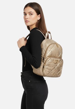 Zaino trapuntato color oro con doppia cerniera, caratterizzato da un motivo con logo del brand e dettagli in metallo color oro, indossato sulla spalla.