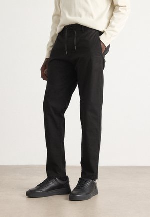 Schwarze Baumwollhosen mit einem elastischen Bund und seitlichen Taschen, kombiniert mit schwarzen Turnschuhen auf einer hellgrauen Fläche.