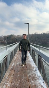 Ponte de madeira com corrimões cinzentos, uma pessoa a caminhar sobre ela. As árvores ao redor estão despidas, e um céu limpo com algumas nuvens espalhadas está ao fundo.