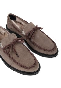 Brązowe zamszowe kapcie z miękkim wnętrzem, ciemną gumową podeszwą oraz charakterystycznym brązowym warkoczowym przeszyciem. Zawierają związane w kokardę sznurówki.