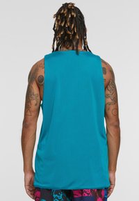 Teal mesh tank top dengan potongan longgar, menampilkan kerah bulat dan desain tanpa lengan; dipadukan dengan celana pendek berpola, menunjukkan tato atletik.