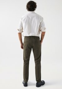 Olijfgroene herenbroek met een slim fit en achterzakken, gecombineerd met een witte lange mouwen shirt. Glad materiaal, rechtlopende pasvorm en zwarte schoenen.