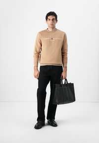 Beige sweatshirt med hvidt og rødt logo, sorte straight-leg jeans og en sort læder tote taske med fire lodrette sømme og dobbelte håndtag.