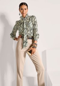 Grüne Bluse mit Schlangenmuster und bindbarem Hals, kombiniert mit beigen, taillierten Hosen. Die Accessoires umfassen Armreifen und eine goldene Clutch.