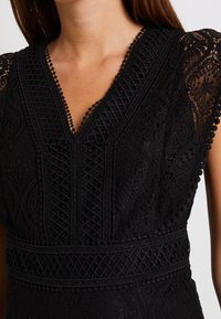 Vêtement en dentelle noire avec un col en V, des motifs complexes et des accents texturés. Il possède des bordures détaillées et divers superpositions de dentelle.