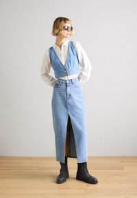 Denimowa krótka kamizelka na białej koszuli z guzikami, w komplecie z długą spódnicą z denimu z rozcięciem z przodu i czarnymi botkami na obcasie.