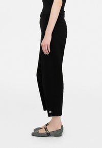 Pantaloni negri cu talie înaltă, dotati cu buzunare laterale și tivuri răsucite, având butoane contrastante la talie. Purtați cu balerini negri cu model.