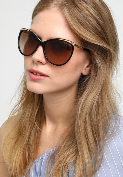 Černé oversized sluneční brýle s gradientními hnědými skly, zlatými akcenty na nohách a lesklým povrchem. Modelka má dlouhé blond vlasy a na sobě modrý top.