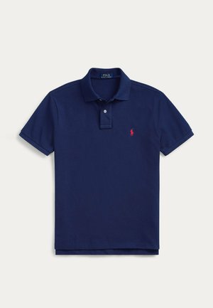 Uomo in piedi e sorridente, con una polo blu navy, jeans blu e sneakers bianche su uno sfondo chiaro e uniforme.