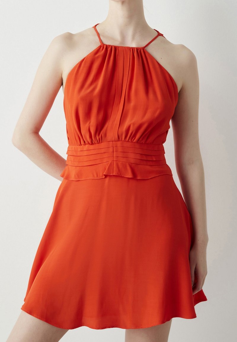 Femme portant une robe sans manches rouge vif à col halter avec des détails plissés à la taille, debout devant un fond clair uni.
