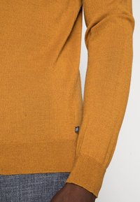 Hořčicově žlutý svetr z měkké pleteniny, s vroubkovaným lemem a manžetami a malou značkovou štítkem na spodní straně.